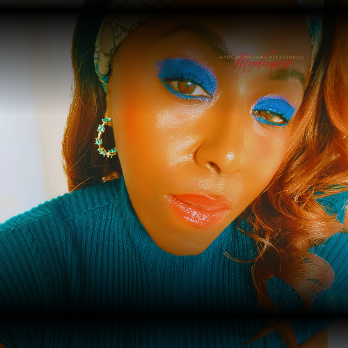 Close-up of a person with blue eyeshadow and orange skin tone wearing a teal sweater.