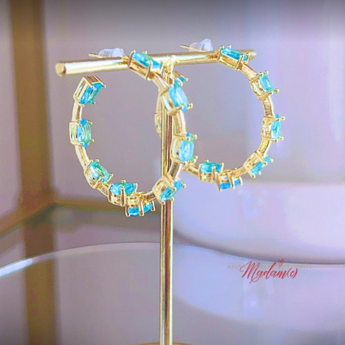Gold hoop earrings with blue gemstones on a stand against a neutral background