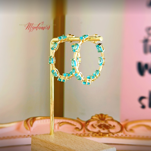 Gold hoop earrings with rectangular turquoise-blue rhinestones, displayed on a small wooden stand against a softly blurred pink and cream background.