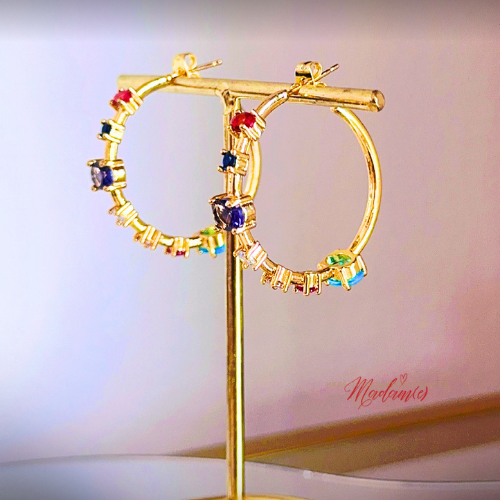 Gold hoop earrings with colorful gemstones on a stand against a light background