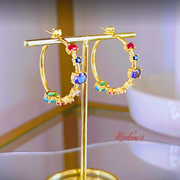 Gold hoop earrings with colorful gemstones on a gold stand, brand 'Madam's' visible.