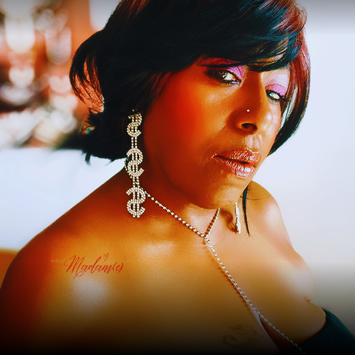 A close-up portrait of a woman with short dark hair, wearing dangling silver earrings and a delicate necklace. The photo has a warm, stylized color cast and a blurred light background.