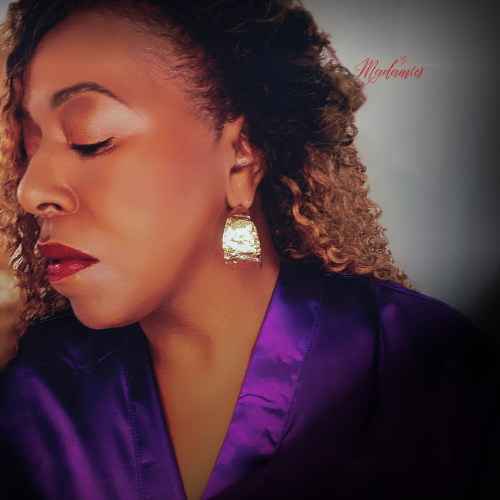 A close-up portrait of a woman with curly hair wearing a shiny purple satin top and a textured gold earring, shown against a dark gray background with a vignette effect.