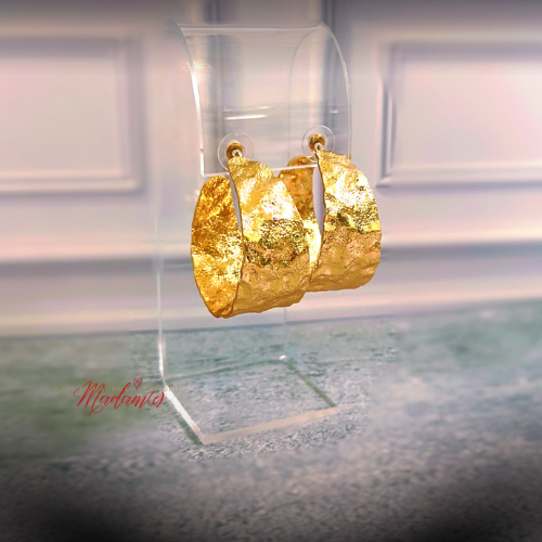 A pair of chunky textured gold hoop earrings displayed on a clear acrylic stand against a softly blurred indoor background.