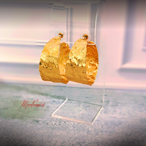 A pair of chunky textured gold hoop earrings displayed on a clear acrylic stand against a softly blurred indoor background.