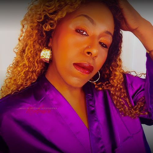 A close-up portrait of a woman with curly blonde hair wearing a shiny purple satin robe and large gold earrings, with a warm orange-toned filter over the photo.