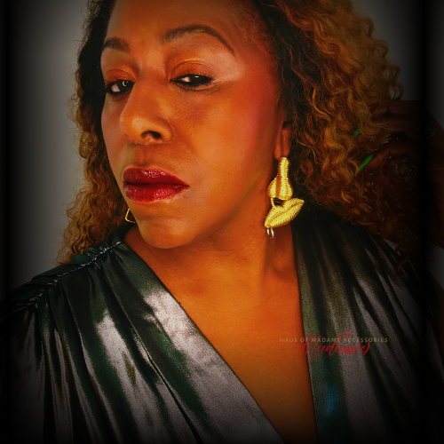 A close-up portrait of a woman with curly hair wearing a shiny dark green satin top and textured gold earrings, set against a dark vignette background.