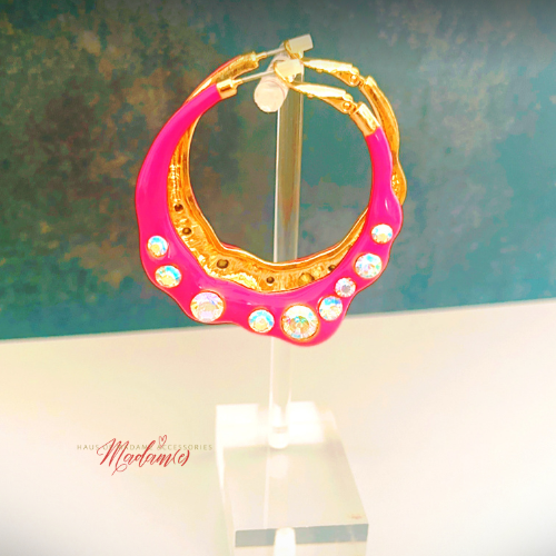 Pink and gold hoop earring with embedded crystals on a stand against a textured green and blue background.