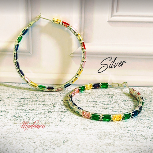 A pair of large silver hoop earrings with rectangular multicolor gemstones, displayed on a light textured surface in front of a white paneled wall. The word 'Silver' is written in black script.